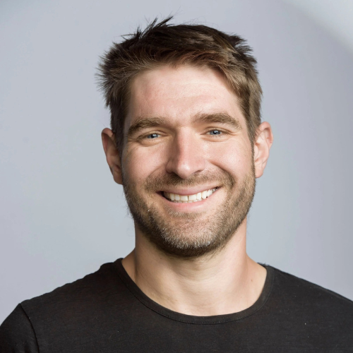 Eben Bayer smiling warmly in a dark shirt against a soft gray studio background.